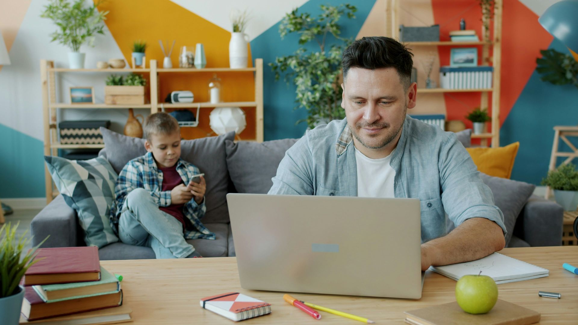 Father works on laptop while son plays on phone.
